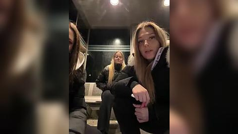 Media: Video of three young women, one with blonde hair, sitting in a modern, dimly-lit room with a minimalist design, wearing dark winter coats, and looking away from the camera.