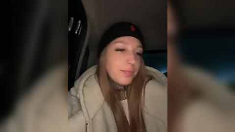 Media: Video of a young Caucasian woman with fair skin, long straight brown hair, wearing a black beanie and a cream-colored puffy jacket, seen through a car window at night, with blurred out-of-focus faces in the foreground.