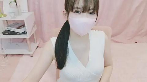 Media: Video of an East Asian woman with long black hair in a ponytail, wearing a white V-neck dress and a pink face mask, seated in a room with pink curtains and a white shelf with books.