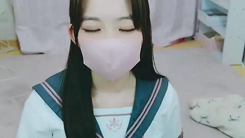 Media: Video of a young Asian girl with long black hair and pale skin, wearing a traditional school uniform with a white blouse and navy blue sailor collar. She has her eyes closed and is wearing a pink face mask. Background shows a pink curtain and a white rug.