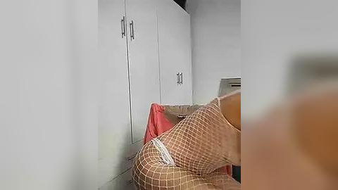 Media: Video of a beige couch with a red blanket and fishnet stockings, set against white cabinets with silver handles, in a minimalist room with light flooring.