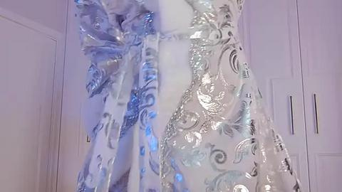 Media: Video of a woman's white gown with shimmering silver and blue floral patterns, worn over a matching sheer blue dress. The background features a white door and wardrobe, emphasizing the elegant, luxurious fabric.