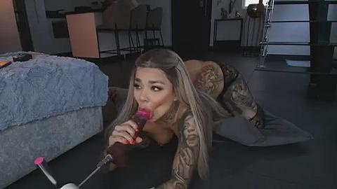 Media: Video of a tattooed woman with long blonde hair, topless, kneeling on a bed, sucking on a pink vibrator, in a dimly lit, modern bedroom.