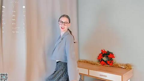 Media: A video of a smiling, fair-skinned woman with glasses, wearing a gray blazer and plaid skirt, stands in front of a green wall. A wooden table with a red floral arrangement and gold garland is visible in the background.