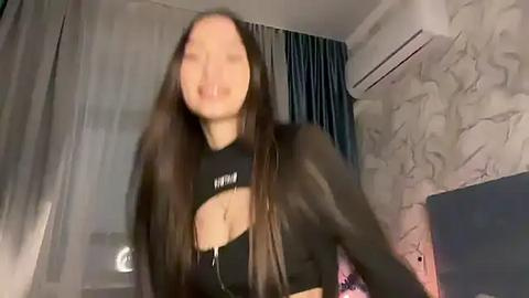 Media: Video of a young Asian woman with long, straight brown hair, wearing a black crop top with cut-outs, standing in a dimly lit bedroom with marble walls and a TV.