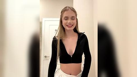 Media: Video of a smiling, fair-skinned blonde woman with long hair in a black zip-up hoodie, white pants, standing in a hallway with beige walls and a white door.