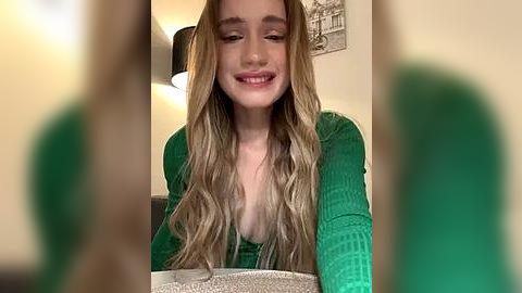 Media: Video of a smiling, light-skinned woman with long, wavy blonde hair, wearing a green ribbed sweater with a low neckline, seated in a warmly lit room with beige walls and a framed black-and-white cityscape.