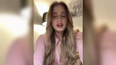 Media: Video of a young woman with long, blonde hair and fair skin, wearing a pink button-up shirt, smiling and leaning forward, in a softly lit room with a black lamp and framed art on the wall.