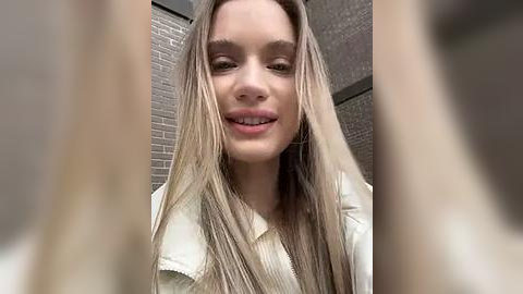 Media: A close-up video of a young woman with long, straight, platinum blonde hair and fair skin, wearing a white ribbed sweater, smiling in a tiled bathroom.