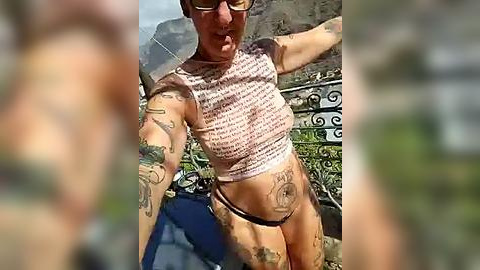 Media: A video of a fit woman with tattoos, wearing a crop top and thong, posing outdoors. Background includes a fence and blurred people.