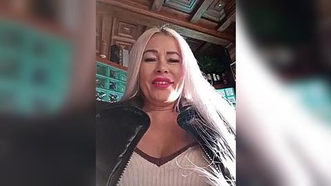 Media: Video of a smiling, light-skinned, blonde-haired woman with long hair, wearing a black jacket over a white top, in a dimly lit, rustic bar with wooden beams and green tiles.