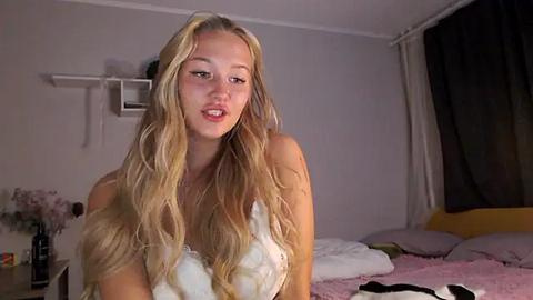 Media: A video of a blonde woman with wavy hair, wearing a white lace bra, sitting on a bed in a dimly lit room. The background features a white wall, a dark curtain, and a white shelf with a few items.