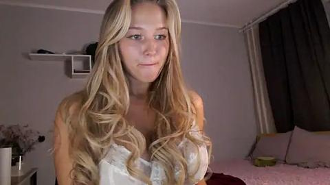 Media: Video of a young blonde woman with long, wavy hair, wearing a white lace bra, standing in a dimly lit bedroom with white walls and dark curtains.