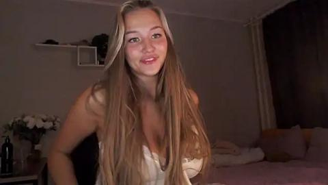 Media: Video of a smiling young woman with long blonde hair, wearing a low-cut white top, standing in a dimly lit bedroom with a bed, flowers, and a window with closed curtains.