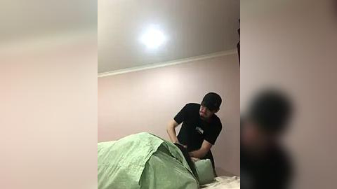 Media: Video of a young man with short black hair in a black t-shirt, standing in a dimly lit, pale pink-walled bedroom with a white ceiling light and a green blanket covering a bed.