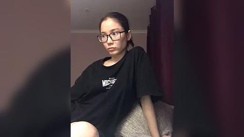 Media: Video of a young woman with fair skin, dark hair, and glasses, wearing a loose black t-shirt, seated on a couch, indoors with maroon curtains.