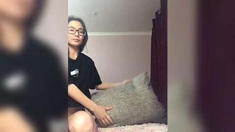 Media: Video of a young Asian woman with glasses, sitting on a bed, wearing a black t-shirt, with a gray pillow beside her, in a softly lit room.