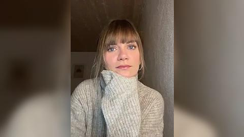 Media: Video of a young woman with fair skin, blue eyes, and blonde hair in a gray sweater, standing in a dimly lit room with a textured wall and blurred background.