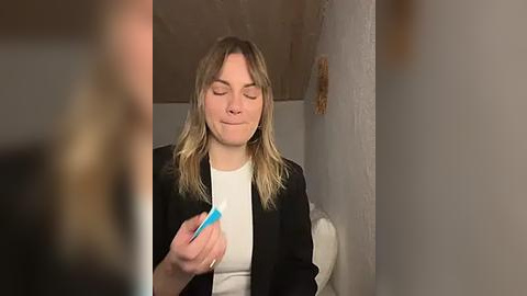 Media: Video of a blonde woman with shoulder-length hair, wearing a white top and black cardigan, holding a blue toothbrush in her right hand, smiling, standing in a dimly lit room with beige walls.