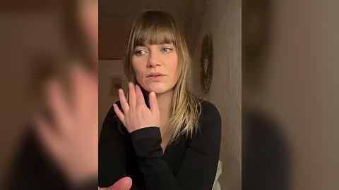 Media: Video of a young woman with straight, light brown hair and bangs, wearing a black long-sleeve top, in a dimly lit, rustic room with a textured wall and indistinct background.