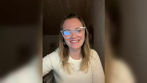 Media: Video of a smiling Caucasian woman with long blonde hair, wearing clear glasses, a white long-sleeve shirt, and a neutral background.