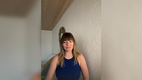 Media: Video of a Caucasian woman with shoulder-length blonde hair, wearing a sleeveless navy blue top, standing in a beige-walled room with wooden ceiling beams and a textured wall.