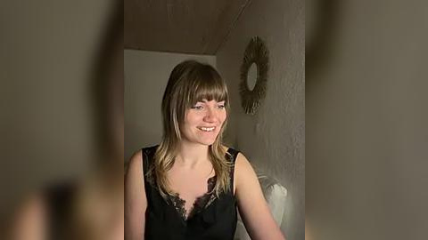 Media: Video of a smiling woman with straight, shoulder-length brown hair, wearing a black lace-trimmed sleeveless top, standing in a dimly lit room with a beige wall and a circular mirror.