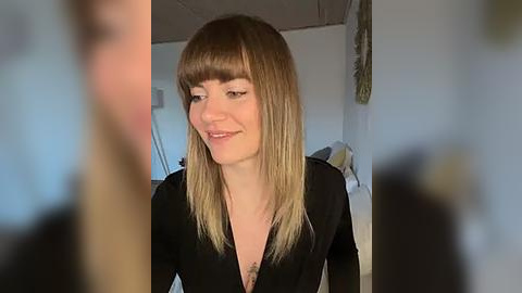 Media: Video of a smiling young woman with shoulder-length blonde hair and bangs, wearing a black dress, indoors, blurry background.