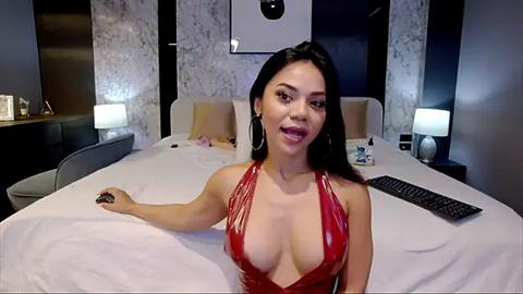 Media: Video of a curvy, light-skinned woman with long black hair, wearing a red, shiny, halter-neck dress, sitting on a bed in a modern, dimly-lit bedroom with white bedding, a keyboard, and a framed photo on the wall.