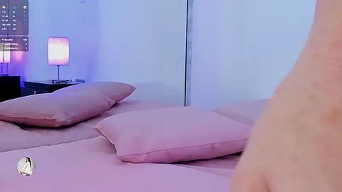 Media: A video of a minimalist bedroom with pale pink bedding, a matching pillow, and a bedside table with a lamp casting purple light. The background features a large mirror and a cityscape view through a window.