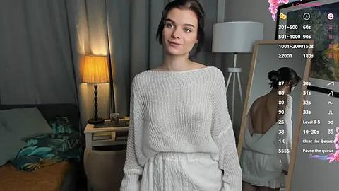 Media: Video of a young woman with short dark hair, wearing a white knitted sweater and high-waisted pants, standing in a modern bedroom with a lamp, mirror, and a digital camera displaying an overlay of camera settings.