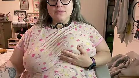 Media: Video of a plus-sized woman with long, grey hair, wearing a pink bunny-themed t-shirt, large black-framed glasses, and a black choker, sitting on a bed in a cluttered bedroom.