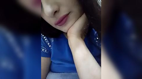 Media: A video of an Asian woman with fair skin, resting her chin on her hand, wearing a blue blouse with white sequins, in a dimly lit room.