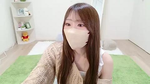 Media: Video of a young Asian woman with long brown hair and light skin, wearing a beige knitted cardigan, white top, and face mask, sitting on a green rug in a minimalist, light-colored room with shelves and toys in the background.