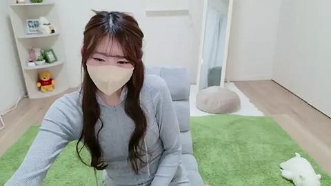 Media: Video of a young Asian woman with long brown hair and a white face mask, wearing a light grey sweater, sitting on a green rug in a minimalist, bright room with shelves, a teddy bear, and a plush chair.