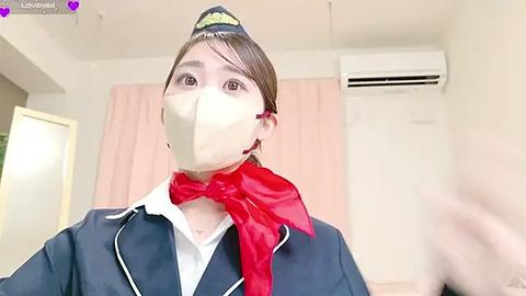 Media: Video of a young Asian woman in a flight attendant uniform with a red scarf, wearing a face mask, standing indoors with pale walls and pink curtains.