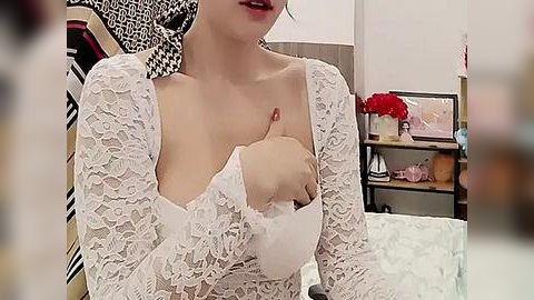 Media: Video of a fair-skinned woman with red lipstick, wearing a white lace dress, sitting on a bed, giving a thumbs-up. Background features a modern, minimalist room with a floral arrangement and a bookshelf.