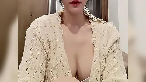 Media: Video of a fair-skinned woman with dark hair, wearing an open cream-colored, knitted cardigan, exposing her cleavage, sitting indoors with a wooden door in the background.