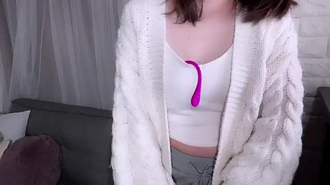 Media: Video of a young woman in a white knit cardigan, revealing a white crop top with a pink tie, and gray sweatpants, leaning against a gray sofa.