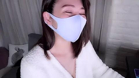 Media: Video of a young woman with light skin and straight brown hair, wearing a light blue surgical mask and a white fluffy bathrobe, smiling with closed eyes in a modern, softly lit bathroom with a gray sink and towel rack.