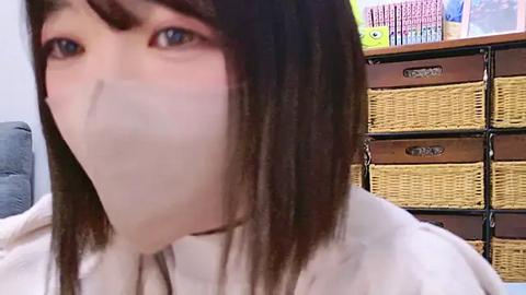 Media: A video of a woman with straight, shoulder-length brown hair, wearing a white surgical mask, sitting in an office with wooden drawers and a wicker basket in the background.