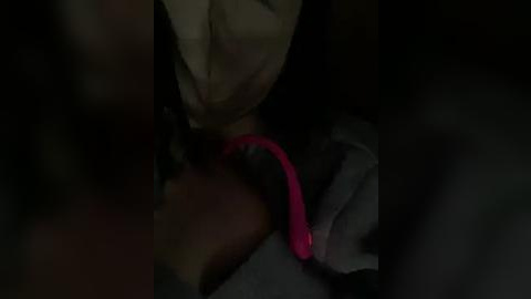 Media: A dimly lit video captures a person's hand holding a bright pink vibrator against a dark background. The individual's beige fabric is partially visible. The scene is intimate and suggestive.