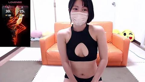 Media: Video of a slim East Asian woman with short black hair, wearing a black sports bra and black panties, seated on a mat in a brightly lit room with a pink heart cushion and orange chair.