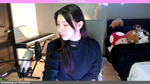 Media: Video of an Asian woman with long black hair, wearing a black turtleneck, sitting at a desk in a cluttered room. Background includes a keyboard, a stuffed animal, and a mirror.