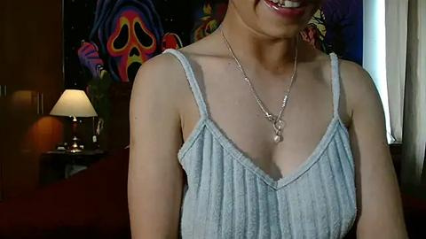 Media: Video of a smiling Asian woman with light skin, wearing a light blue, ruffled tank top and silver necklace, in a dimly-lit room with vibrant wall art, a lamp, and a red couch in the background.