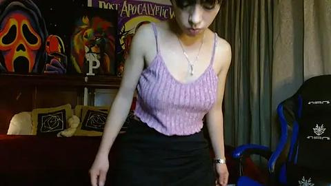 Media: Video of a young woman with light skin and brown hair, wearing a purple ribbed tank top and black skirt, standing in a room with colorful posters, a gaming chair, and dark curtains.