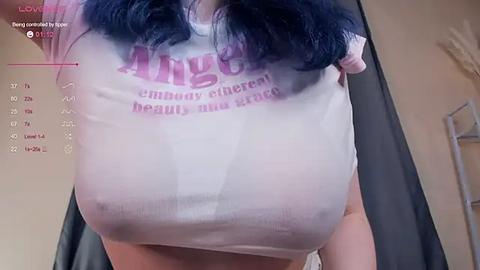 Media: Video of a woman with long, dark blue hair wearing a wet, white t-shirt with \"Angel\" printed in pink. Her large, bare breasts are visible through the shirt.