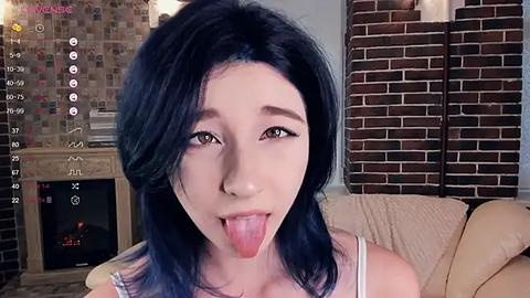 Media: Video of a young Asian woman with blue hair, sticking out her tongue, in a cozy living room with a brick fireplace and a beige couch.