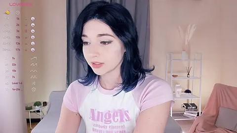 Media: Video of a young woman with pale skin and shoulder-length blue hair, wearing a light pink Angels t-shirt, seated in a minimalist room with a white shelving unit, a potted plant, and a pink blanket.