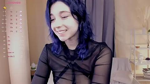 Media: Video of a young, light-skinned woman with blue hair, wearing a sheer black top, smiling in a bedroom with a calendar, bed, and shelving unit in the background.